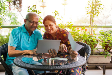 Senior Indian/asian couple accounting, doing home finance and checking bills with laptop, calculator and money while sitting on sofa/couch at home
