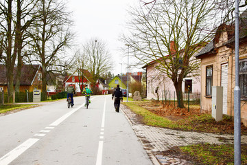 Two teen boys riding bicycles on street landscape with residential houses and naked trees in early spring day, man walking on pedestrian path, back view.