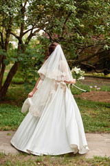 Fototapeta na wymiar Beautiful bride in long wedding dress and veil with bridal bouquet walking in park and waiting for groom before wedding ceremony, back view, copy space. Bride morning