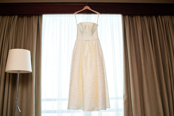 Beautiful beige embroidered wedding dress hanging on hanger against window in the room, copy space. Bridal morning preparations