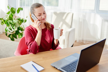 Happy young business woman sitting at wood desk, enjoying coffee and talking on cellphone in modern office, copy space