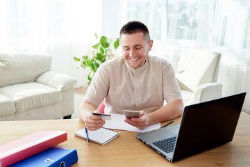 Happy cheerful man paying with credit card on smart phone at home office, copy space. Technology, banking, business, online shopping concept