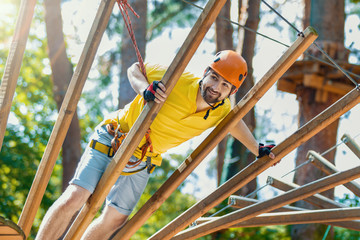 Young male man adult wears protective helmet with action camera having fun in extreme rope park, amusement park. Climbing in rope bridge at green forest. Active healthy lifestyle in spring or summer.