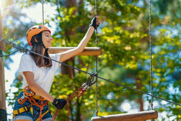 Young beauty female woman adult wears protective helmet having fun in extreme rope park, amusement park. Climbing in rope bridge at green nature forest. Active healthy lifestyle in spring or summer.