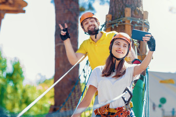 Young woman and man in protective gear are standing on rope bridge hanging on high trees, making selfie and smiling. Rope park with obstacles and ziplines. Extreme rest and summer activities concept.
