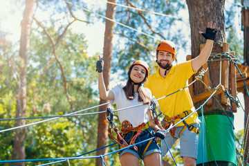 Young woman and man in protective gear are standing on rope bridge hanging on high trees, posing and smiling. Rope park with obstacles and ziplines. Extreme rest and summer activities concept.