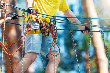 Young male man adult wears protective helmet with action camera having fun in extreme rope park, amusement park. Climbing in rope bridge at green forest. Active healthy lifestyle in spring or summer. - Powered by Adobe
