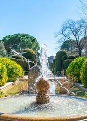 A fountain in the foreground with a sculpture of flying birds over a pool and a path lined with trimmed trees and shrubs in the background.