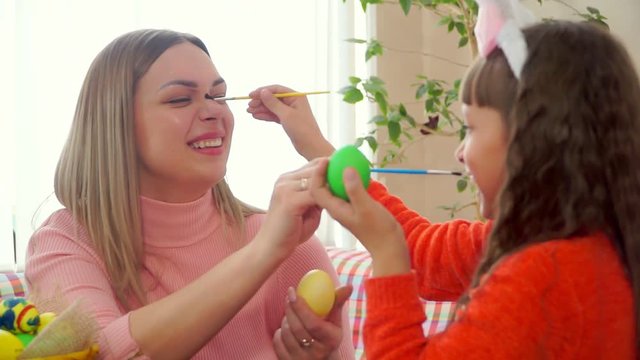 close - up mom and daughter paint Easter eggs, and then brushes tickle each other's noses.