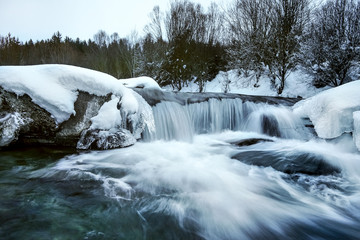 River covered with snow and ice in winter, trees background, long exposure photo with milky smooth water flow.