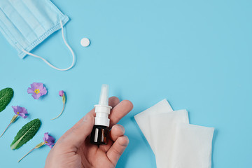 Creative flat lay concept of seasonal spring and summer pollen allergy with napkins, pills, face mask, drops bottle in male hand and flowers on blue background. Top view, copy space, minimal style