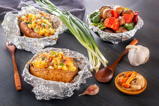 Baked potatoes with bacon, onions and baked vegetables in foil - tomatoes, eggplants, peppers on a gray wooden table.