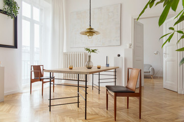 Eclectic and elegant dining room interior with design sharing table, chairs, gold pedant lamp, abstract paintings and stylish accessories. View to living room. Eclectic decor. Brown wooden parquet.