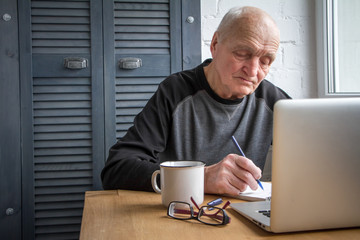 Elderly man working on laptop, looking at screen, makes notes in a notebook and drinking coffee.