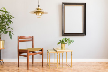Stylish and design retro interior with design chair, gold lamp, small tables with  plant. Black mock up frame on the grey background wall. Minimalistic concept of sitting room. Real photo. 