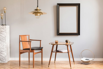 Design retro interior with beautiful chair, gold lamp and small tables. Black mock up frame on the grey background wall. Marble stand with lamp. Minimalistic concept of sitting room. Real photo. 