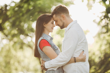 Fototapeta premium Portrait of young romantic couple in love, smiling and embracing in garden. Sweet lovers in casual clothes, posing touching each other face to face, standing in summer park. Sunset time.
