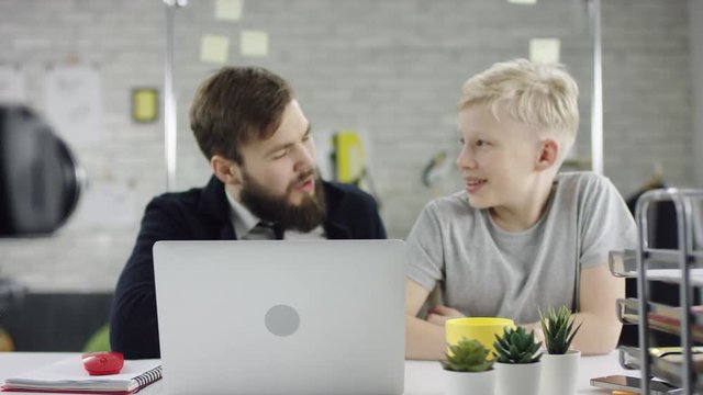 Genius boy wonder hacks system at cyberspace. Cute kid young programmer developer working on computer at office. He is looking in the cam and speaking with a businesswoman