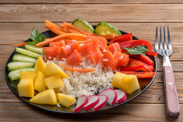 Top view of a Poke salad from hawaii in black plate with rustic fork on wooden background. Close up. Soft focus. To prepare it just take rice, salmon and any vegetables.7