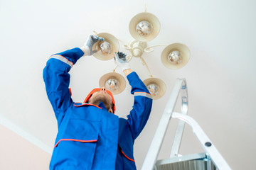 worker in uniform standing on the ladder and repair the chandelier at home f