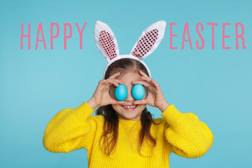 Little girl in bunny ears headband holding Easter eggs near eyes on color background