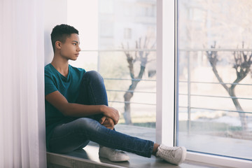 Upset African-American teenage boy sitting alone near window