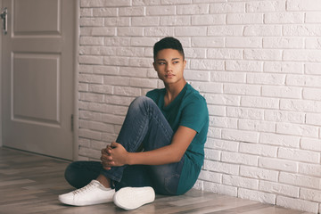 Upset African-American teenage boy sitting alone on floor near wall