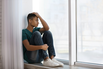 Upset African-American teenage boy sitting alone near window - Powered by Adobe