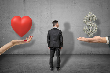 Fototapeta premium Businessman standing between hand holding red heart and hand holding money dollar sign on grey wall background