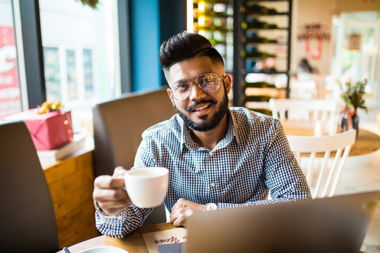 Young Asian Man With Hat Using Laptop At Cafe. Young Indian Man Working On Laptop