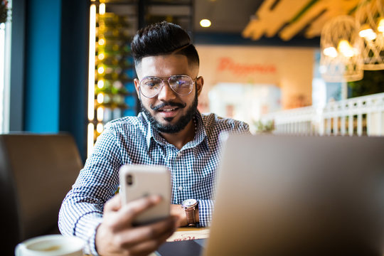 Closeup shot of a young indian man typing a text on mobile phone. Guy holding a modern smartphone and writing a phone message. Smiling young businessman looking at cellphone with laptop on table.