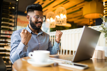 Portrait of handsome successfull indian bearded young freelancer are sitting in cafe and working on laptop win sign
