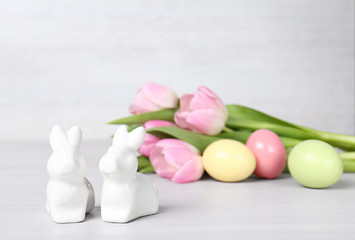 Cute ceramic bunnies, Easter eggs and spring tulips on table against light background, space for text