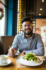 Fototapeta na wymiar Handsome indian bearded man in checked shirt holding fork eating in cafe and smiling looking at camera
