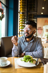 Fototapeta na wymiar Handsome indian bearded man in checked shirt holding fork eating in cafe and smiling looking at camera