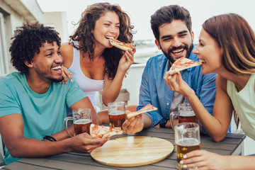 Friends enjoying pizza. Group of young cheerful people eating pizza and drinking beer while sitting at the bean bags on the roof of the building