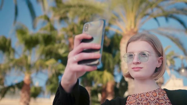 Low shot of young blond attractive and smiling woman in stylish outfit and sunglasses taking selfie with smartphone enjoying holidays on a sea and summer in California background palms blue sky