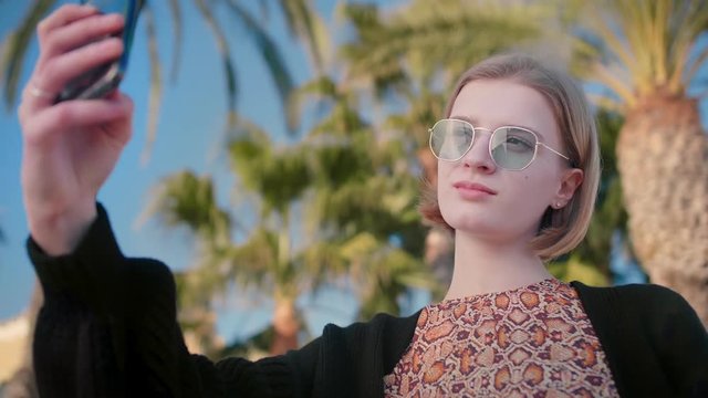 Close up of blond beautiful and smiling woman in trendy outfit and sunglasses taking selfie with smartphone enjoying holidays on a sea, sun and summer background palms blue sky slow motion