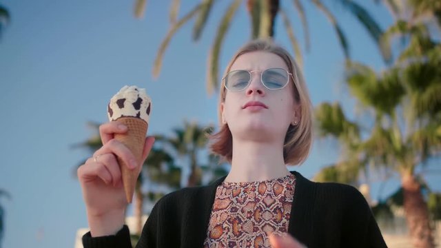 Low shot of blond beautiful woman in trendy outfit and sunglasses eating ice cream, enjoying dessert, sun, beach, holidays on a sea, and summer in California background palms blue sky slow motion