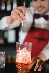 Bartender using long metal spoon to mix ice cubes with drink in a glass standing at wooden bar counter with bottles of alcohol, blurry background.