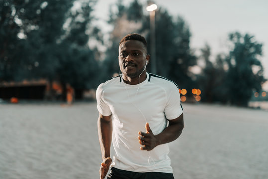 Smiling african male runner in white t-shirt running at the beach. Earphones in ears. One run can change your day, many runs can change your life.