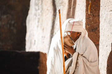Old priest in white robe with walking stick and glasses reading the bible at the lalibela church in...