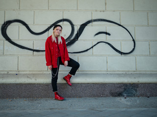 young blonde in a knitted black hat, red jacket and red shoes stands under a bridge amid graffiti