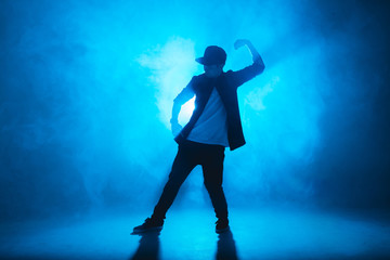 Sporty modern style hip-hop dancer dressed in urban style wear shows his dance on blue studio background.