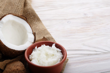 Coconut oil in brown clay bowl and shell with meat on hemp sackcloth on white wooden kitchen table