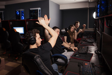 Enthusiastic e-sport gamers. Joyful young people sitting at powerful computers in gaming centre and raising hands triumphantly, celebrating victory in game. - obrazy, fototapety, plakaty