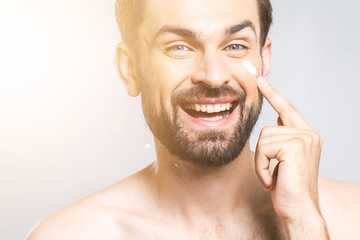 Obraz premium Skin care. Handsome happy young shirtless man applying cream at his face and looking at himself with smile while standing over gray background and looking at camera. Close-Up. Space for text.