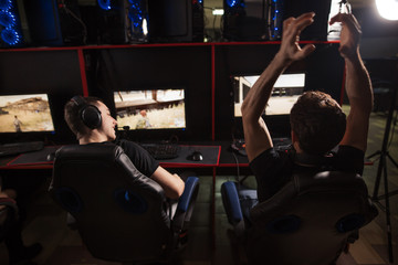 Rear view of focused young gamers sitting in line behind monitors in headsets, playing competitive online game at dark pc gaming club,