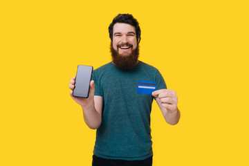Cheerful bearded man looking at the camera showing smart phone and his credit card on yellow background.