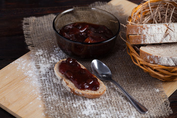 Freshly sliced pieces bread in a wicker bowl on an oak board with scattered flour on a wooden background with of linen fabric next to a bowl of a delicious jam and a spoon